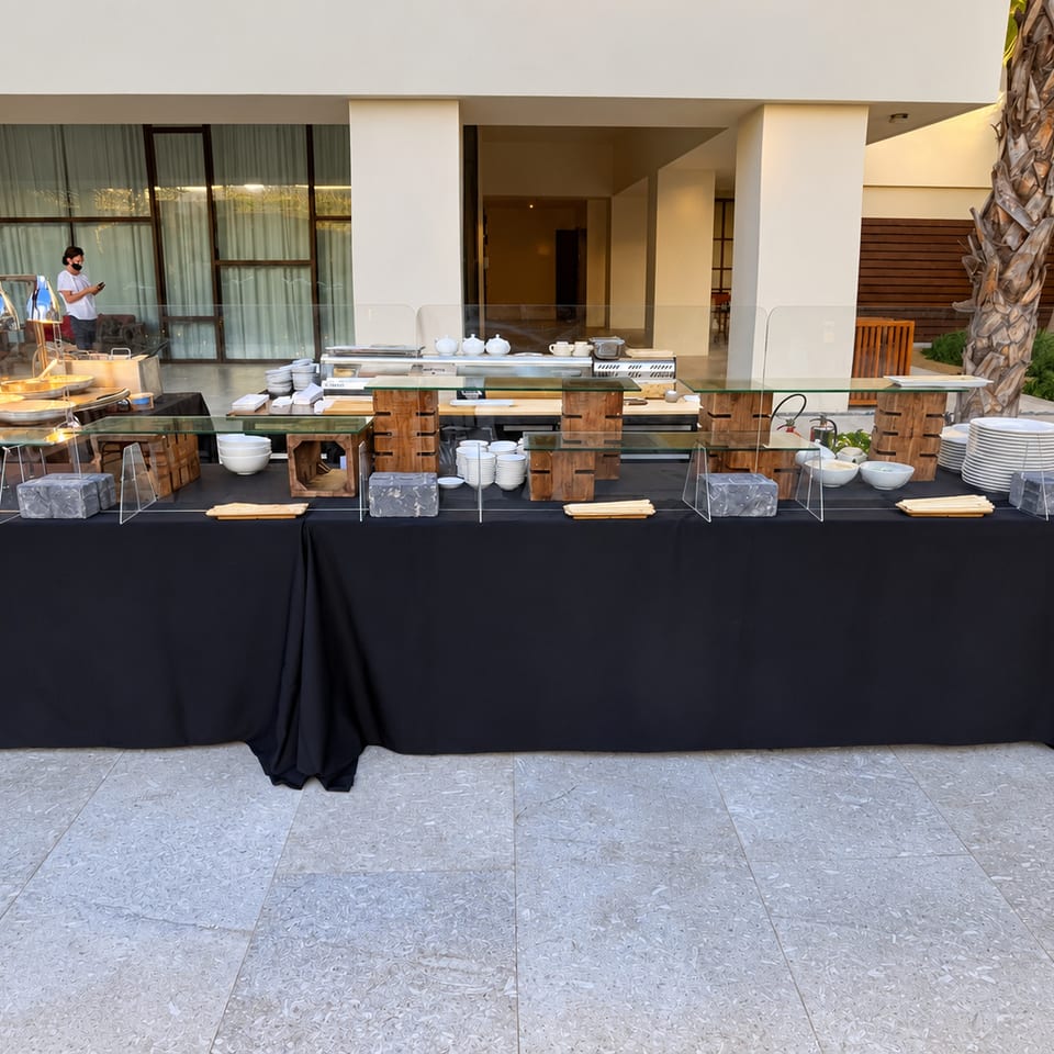 Staffed outdoor buffet with stacked plates and bowls beneath a glass guard