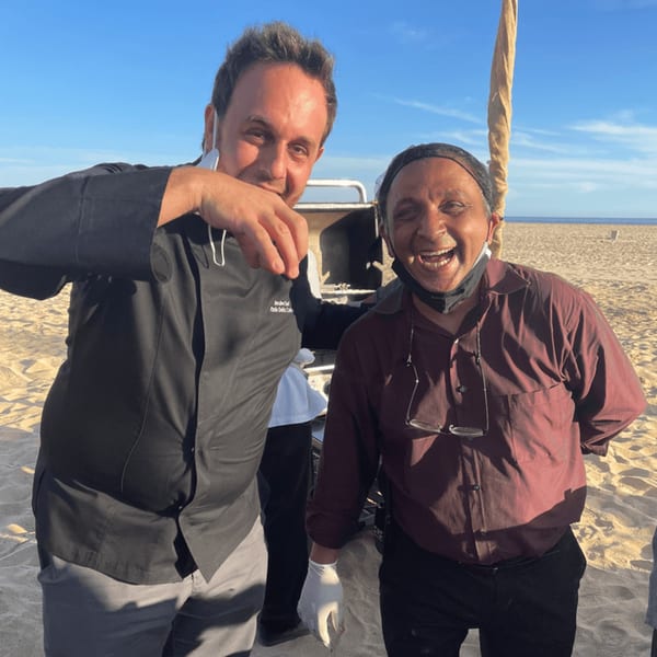 Two men, one in a chef's coat, smiling on a sandy beach under a clear blue sky.