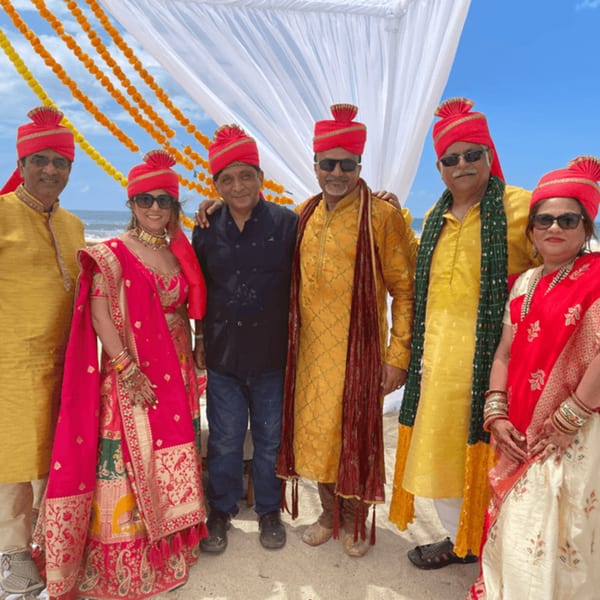 Six people in traditional Indian attire, including red turbans, stand on a beach under a decorated arch.