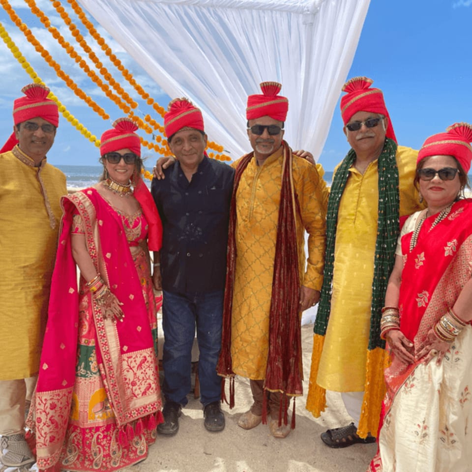 Six people in traditional Indian attire, including red turbans, stand on a beach under a decorated arch.