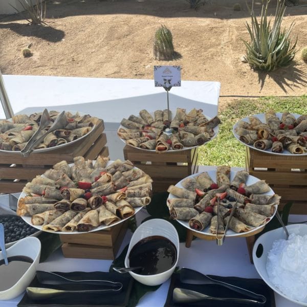 A buffet table with multiple platters of rolled crepes, berries, and dipping sauces outdoors.